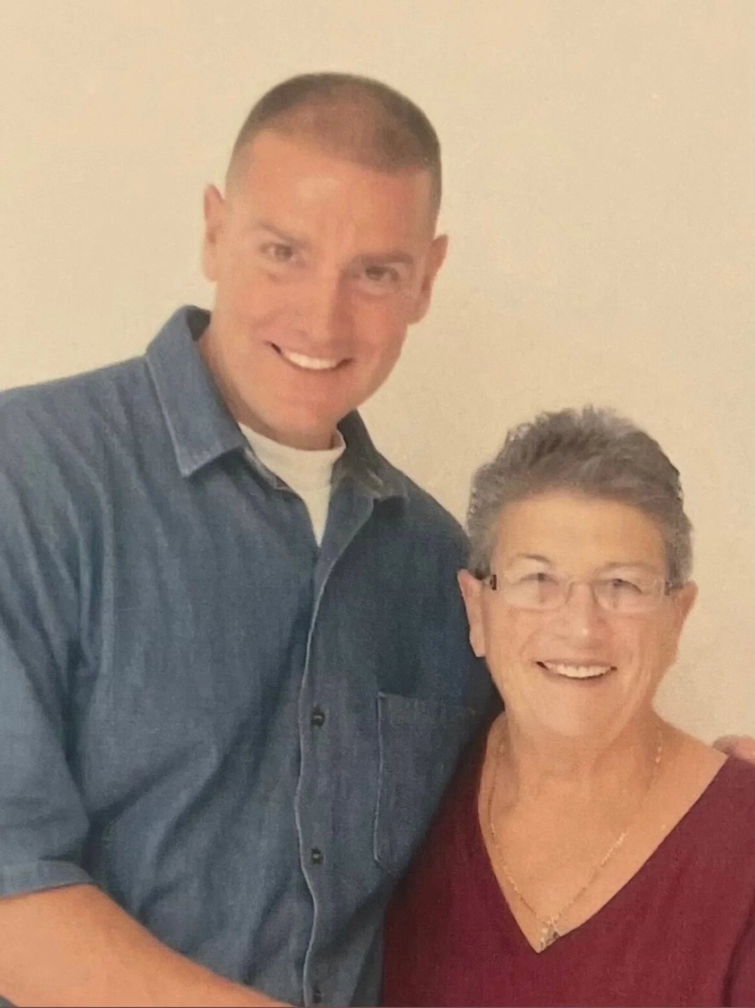 A young man and an older woman smiling for a photo, standing indoors against a plain wall.