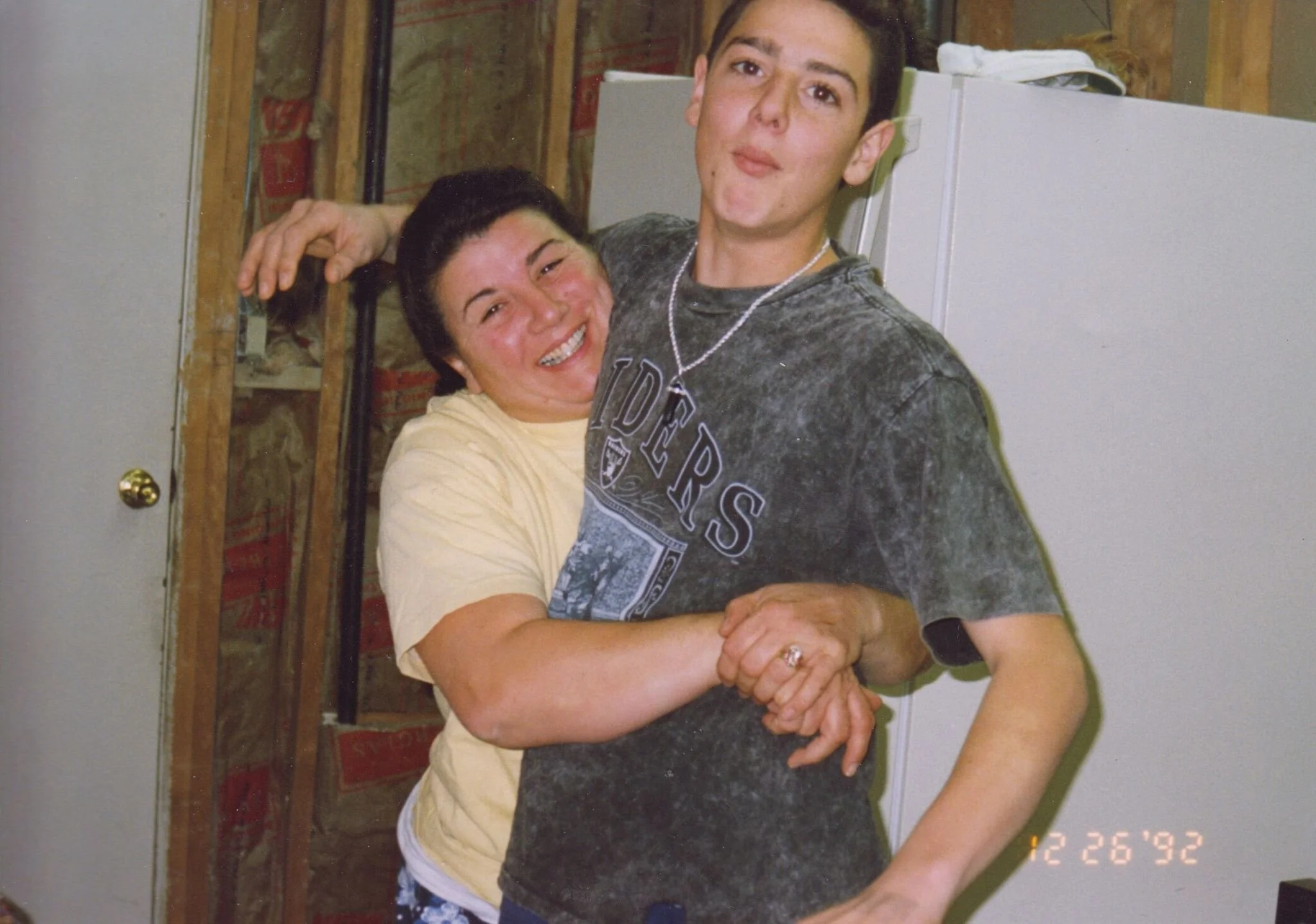A woman and a young man hugging each other in a kitchen, both smiling. The woman has short dark hair and is wearing a yellow shirt, while the young man has short hair and is wearing a dark T-shirt with a design and a necklace. The photo appears vintage, dated December 26, 1992.