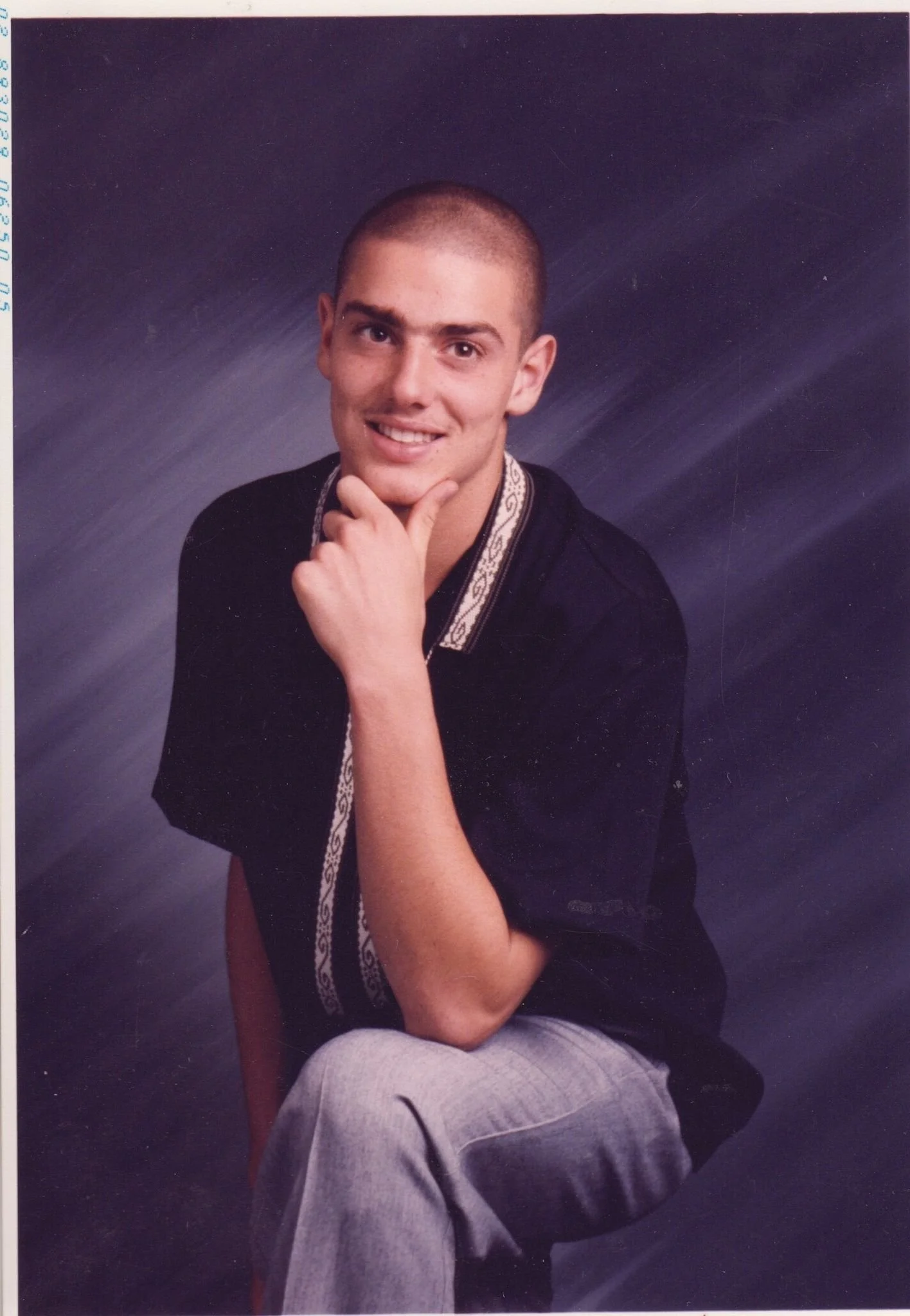 A young man with a shaved head sitting on a stool and posing for a portrait photographer, wearing a black shirt and light gray pants, with a patterned collar detail, against a dark, textured background.