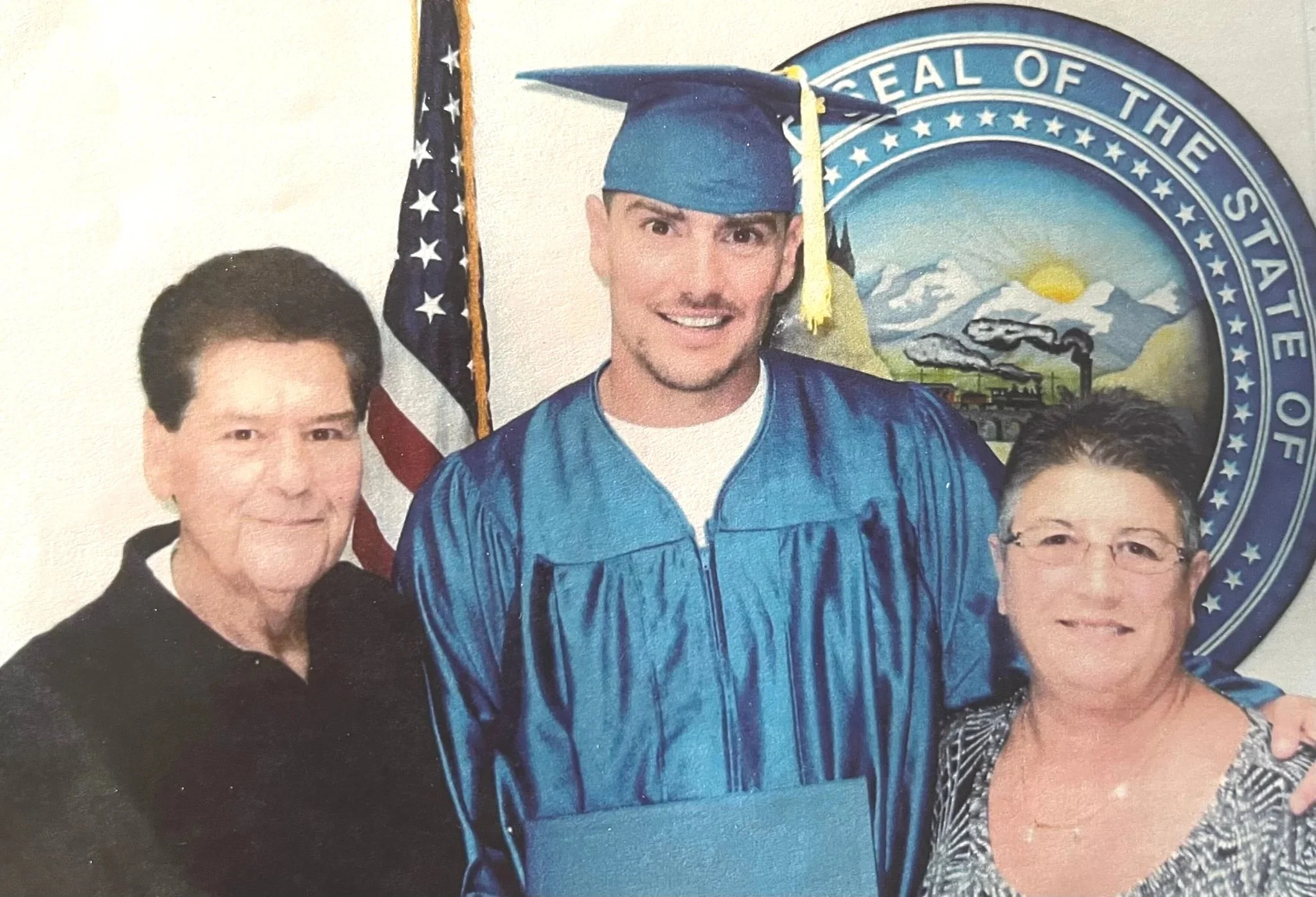A young man in cap and gown standing between an older man and woman, with the state seal of Nevada and American flag in the background.