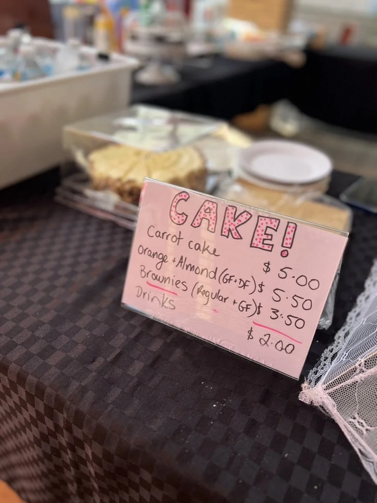 Handwritten sign advertising carrot cake, orange and almond brownies, and drinks at a dessert stand, with a cake and other treats in the background.