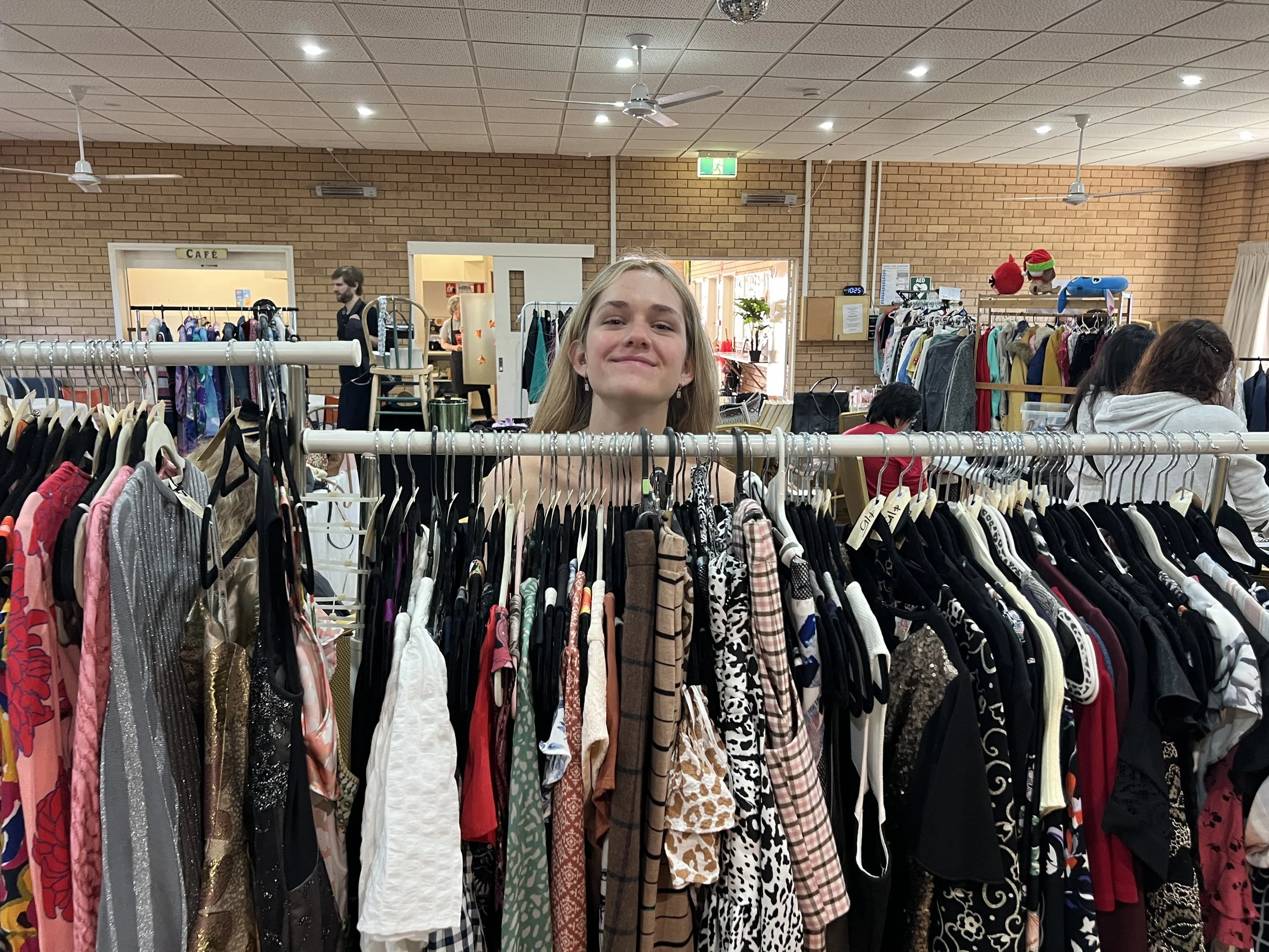 Young woman smiling behind a clothing rack filled with various shirts and dresses inside a store.