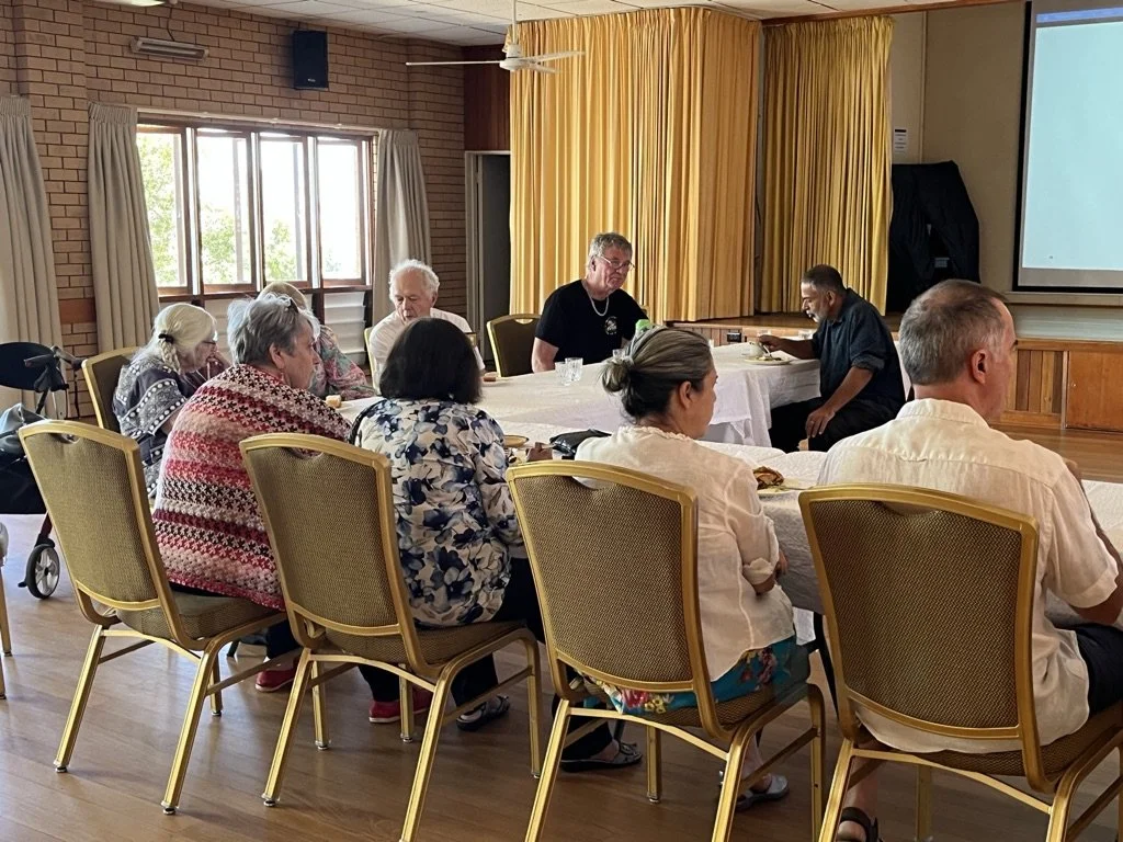 A group of people sitting at a long table in a conference or meeting room, with some eating and listening to a speaker or presentation at the front. A window with curtains and a projector screen are visible in the background.