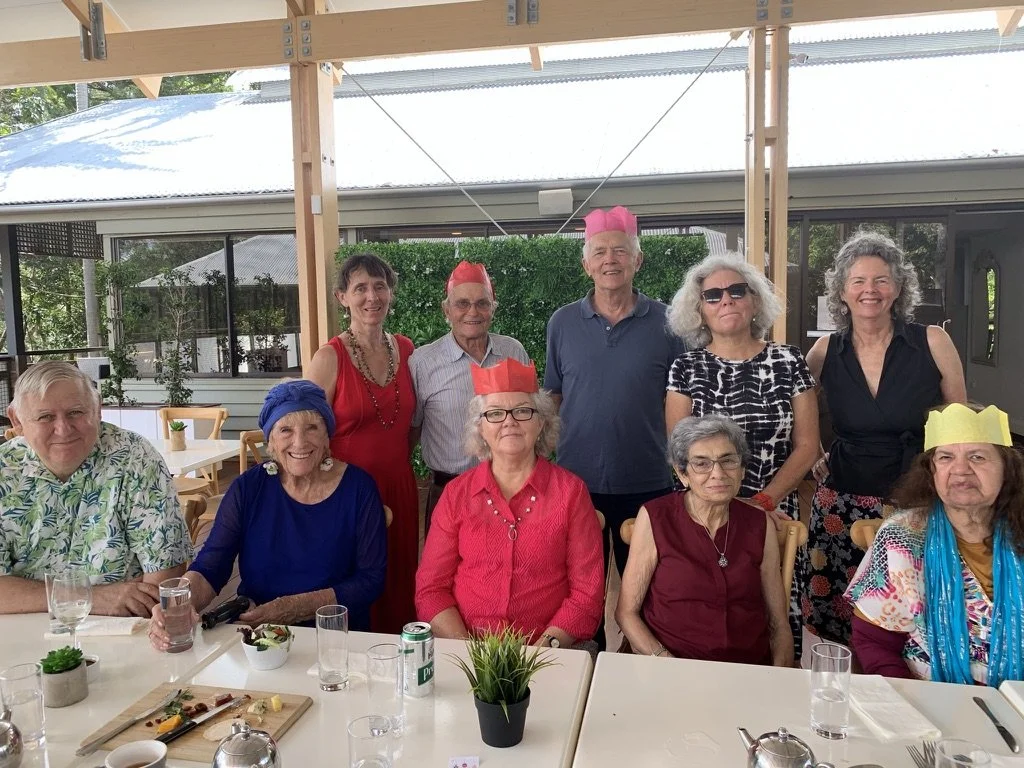 Ten seniors celebrating a party, some wearing colorful paper hats, seated and standing around a table on a patio.