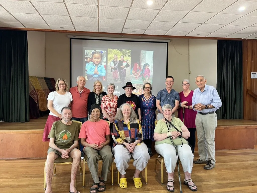 Group of 13 people posing in front of a presentation screen in a room with wooden floors and green curtains. The screen shows photographs of children and people in colorful outdoor settings.
