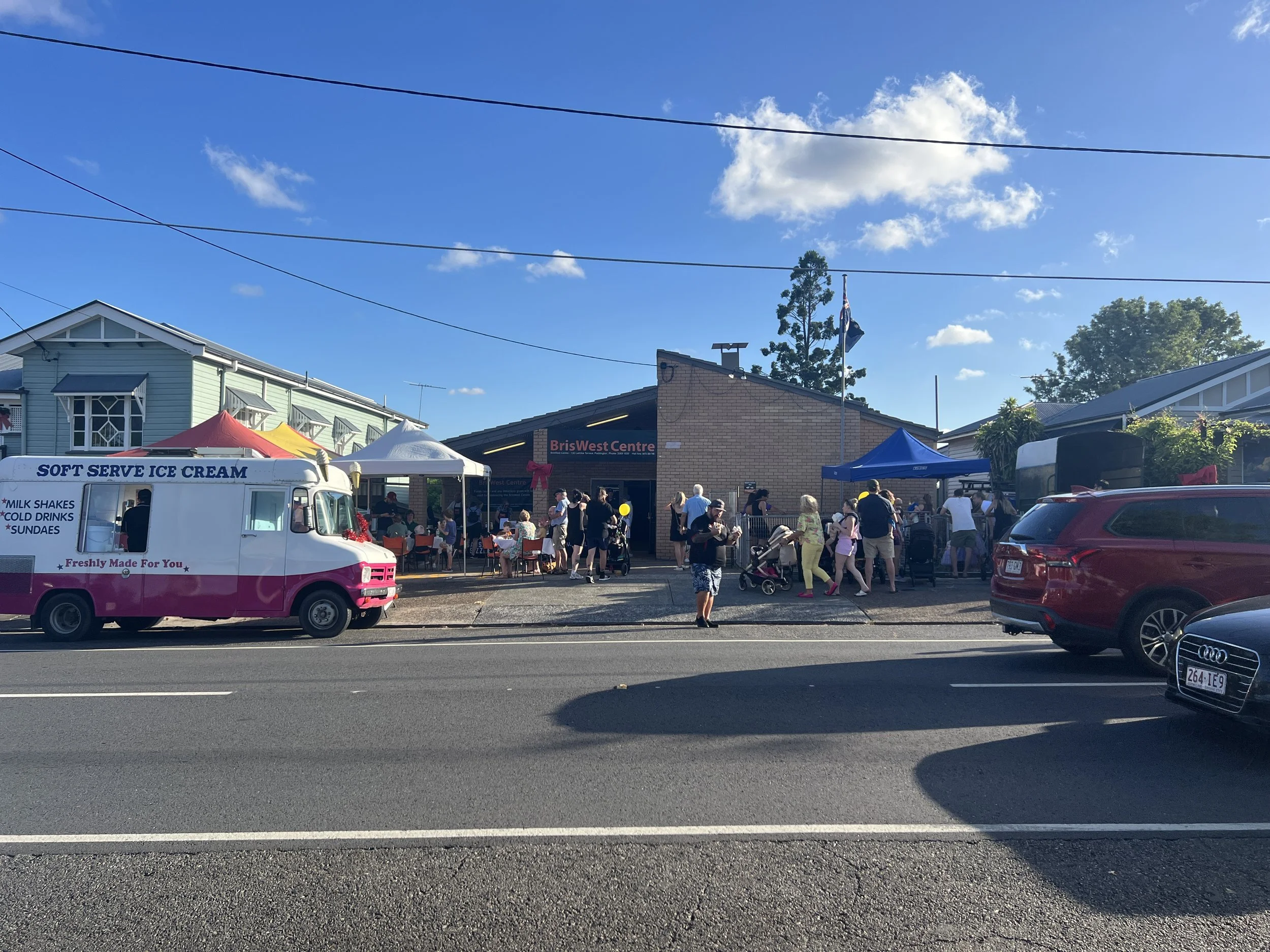A daytime street scene outside BrisWest Centre with people gathered around tents and an ice cream van, cars parked on the road, and a building in the background under a blue sky with some clouds.
