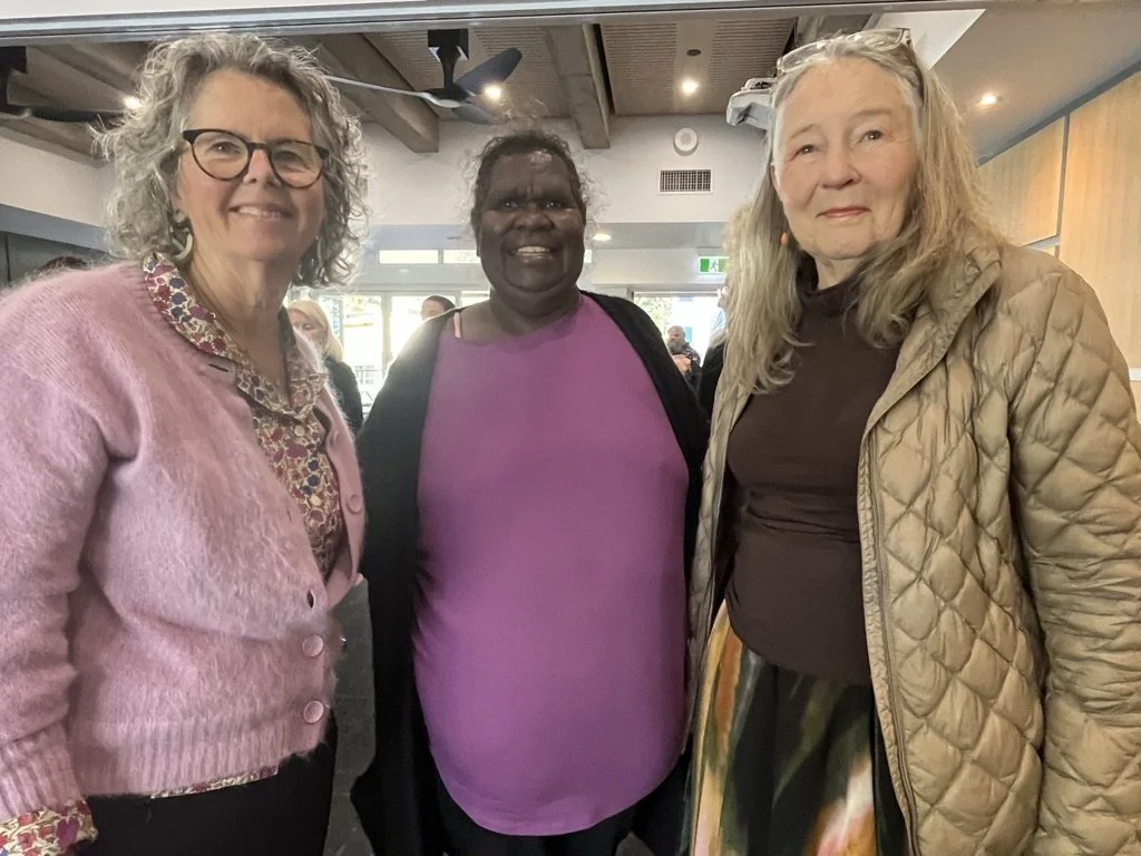 Three women standing together indoors, smiling. The woman on the left has curly gray hair, glasses, and is wearing a pink cardigan over a patterned blouse. The woman in the middle has dark skin, short dark hair, and is wearing a purple top with a bla