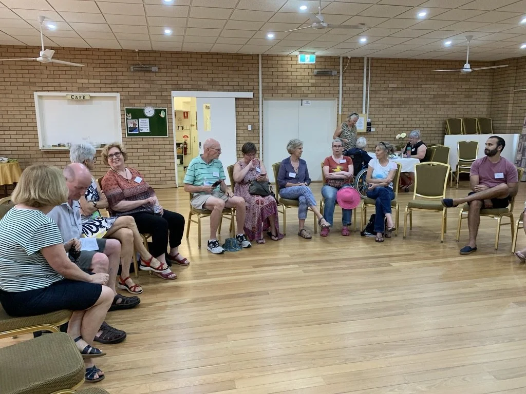A group of elderly and middle-aged people sitting in a semi-circle in a community hall with brick walls, some using walkers, and a woman standing near the wall.