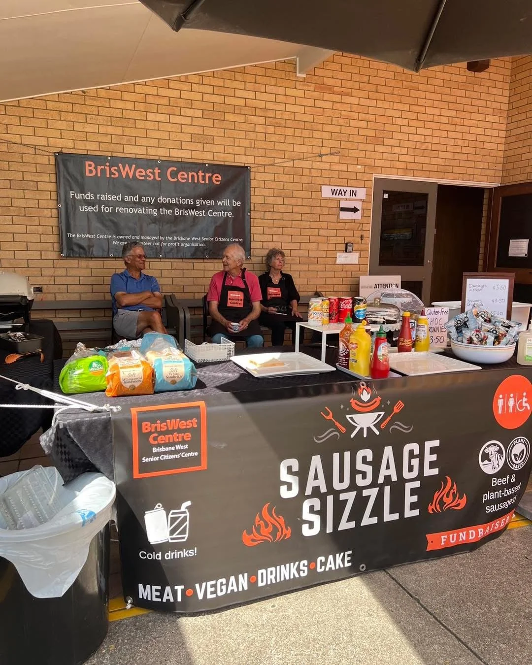 A food stand with a banner and three senior people sitting behind it, selling sausage sizzle and treats at the BrisWest Centre, decorated with various signs, condiments, and snacks.