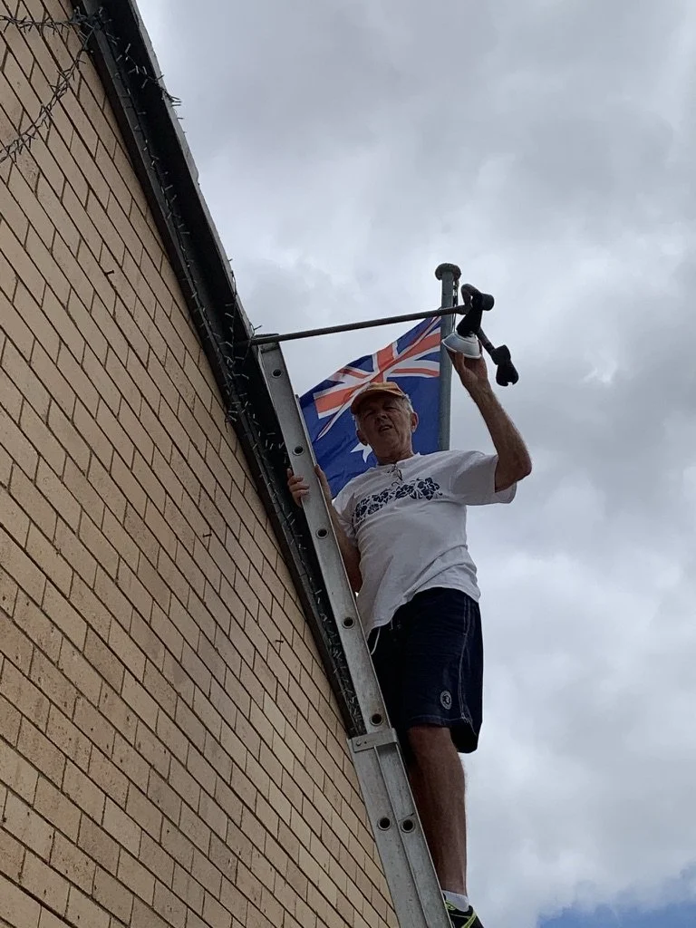 An elderly man standing on a ladder installing a flag on a brick building, with a cloudy sky in the background.