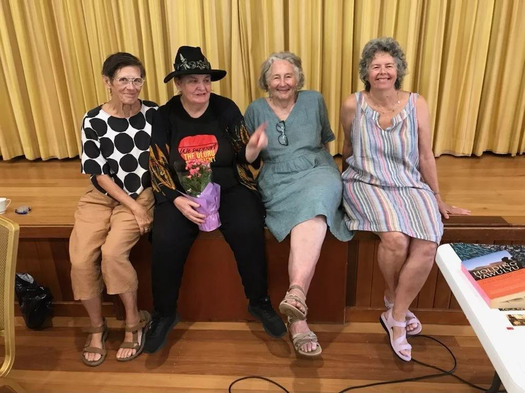 Four women sitting on a stage in front of yellow curtains, smiling for the photo. The second woman from the left is holding a potted plant wrapped in purple paper.