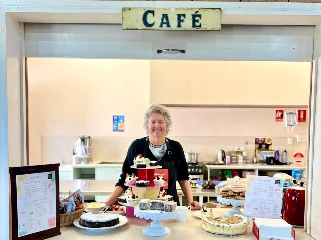 Smiling woman standing behind a counter with various desserts, cupcakes, and pastries at a cafe.