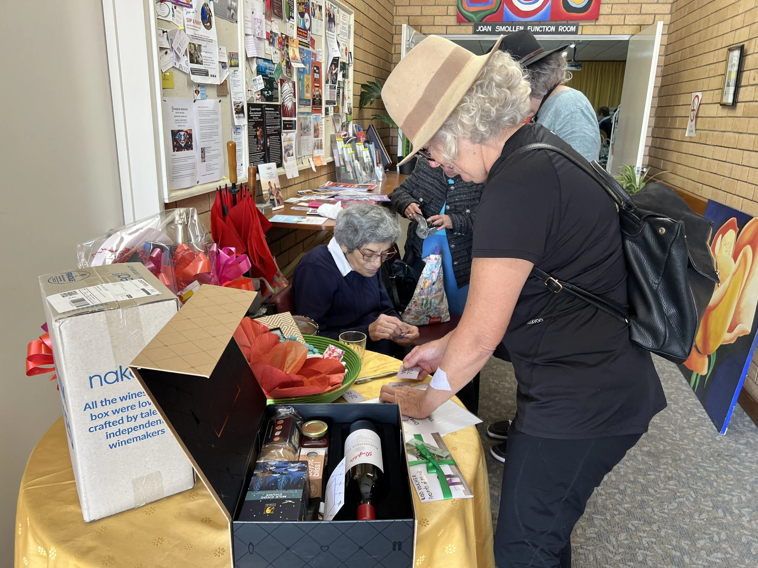 A woman with curly gray hair wearing a wide-brimmed hat looks at items on a table at a fundraising or charity event, with an older woman sitting behind the table. The table has various items, including a gift box with a wine bottle, wrapped gifts, an
