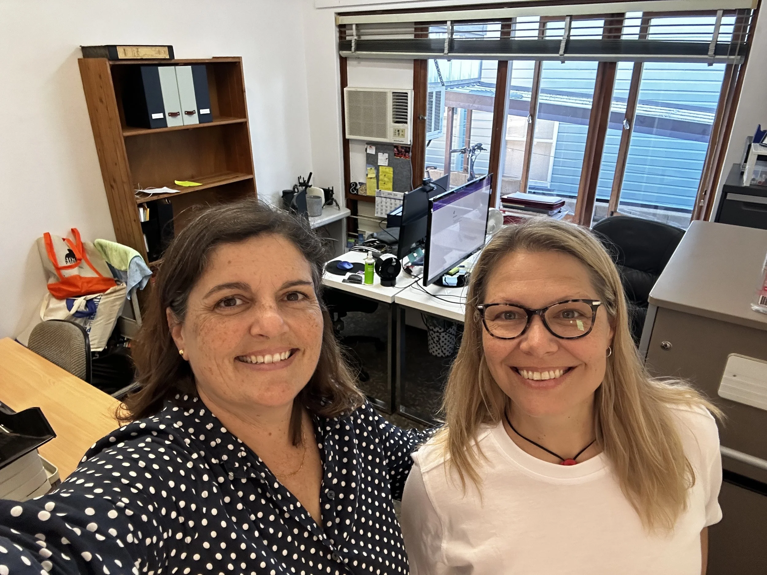 Two women smiling for a selfie in an office. One woman has dark hair and is wearing a black polka-dot shirt. The other woman has blonde hair, glasses, and is wearing a white shirt.