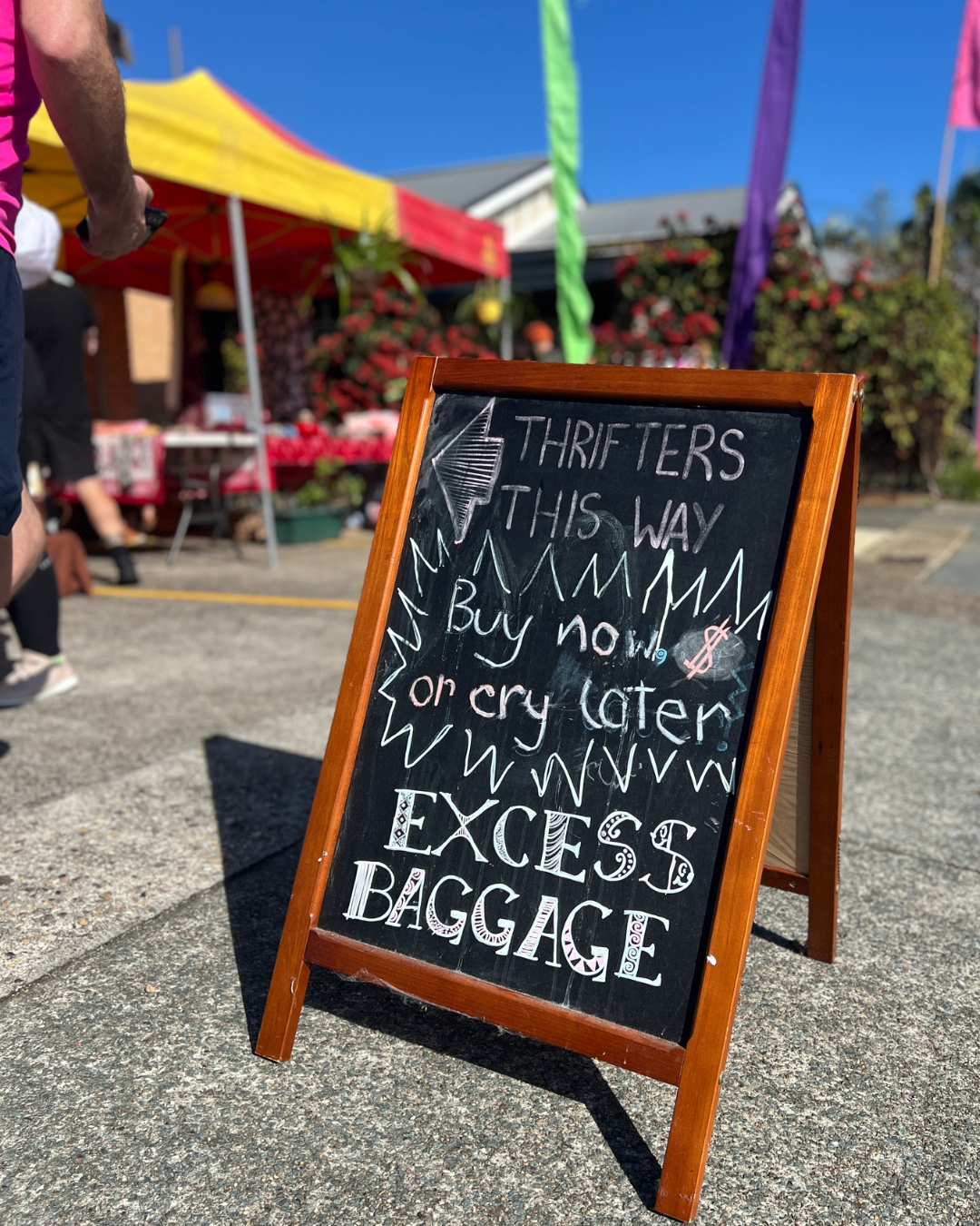 A chalkboard sign at a market with colorful tents in the background, indicating the direction for thrifters to buy or donate!