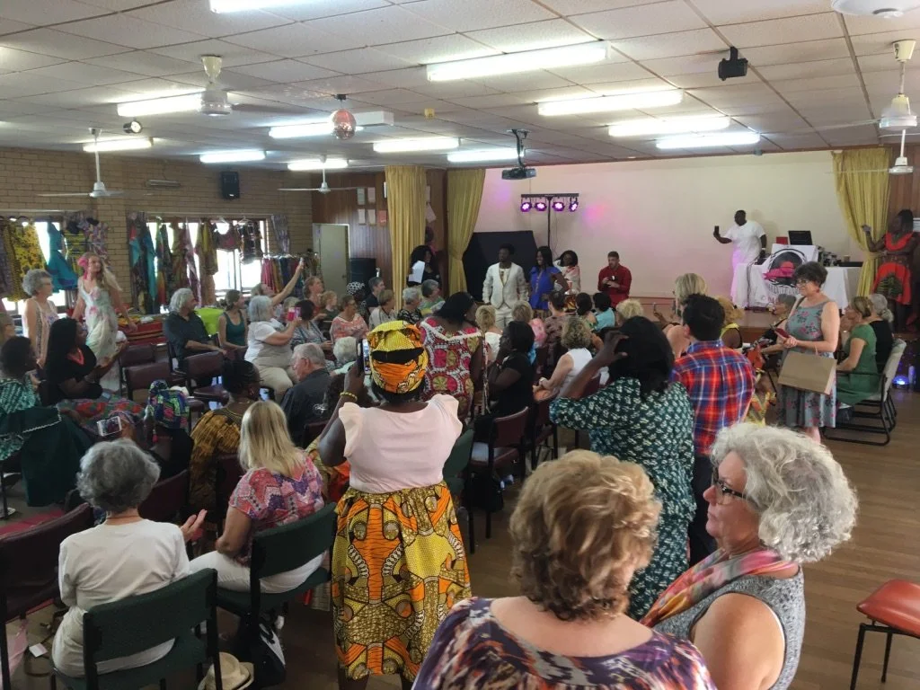 People gathered in a hall, many standing and some sitting, attending a cultural event or celebration with performers on stage, colorful clothing, and decorations.
