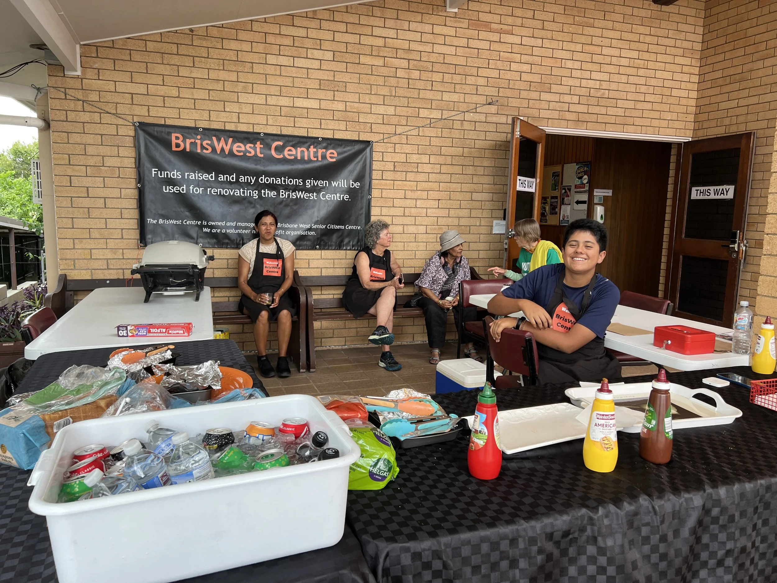 A group of people sitting and smiling inside a community center with a food preparation table in the foreground. The table has condiments, bottled water, and snacks. A banner reads 'BrisWest Centre' and mentions donations for renovating the center.