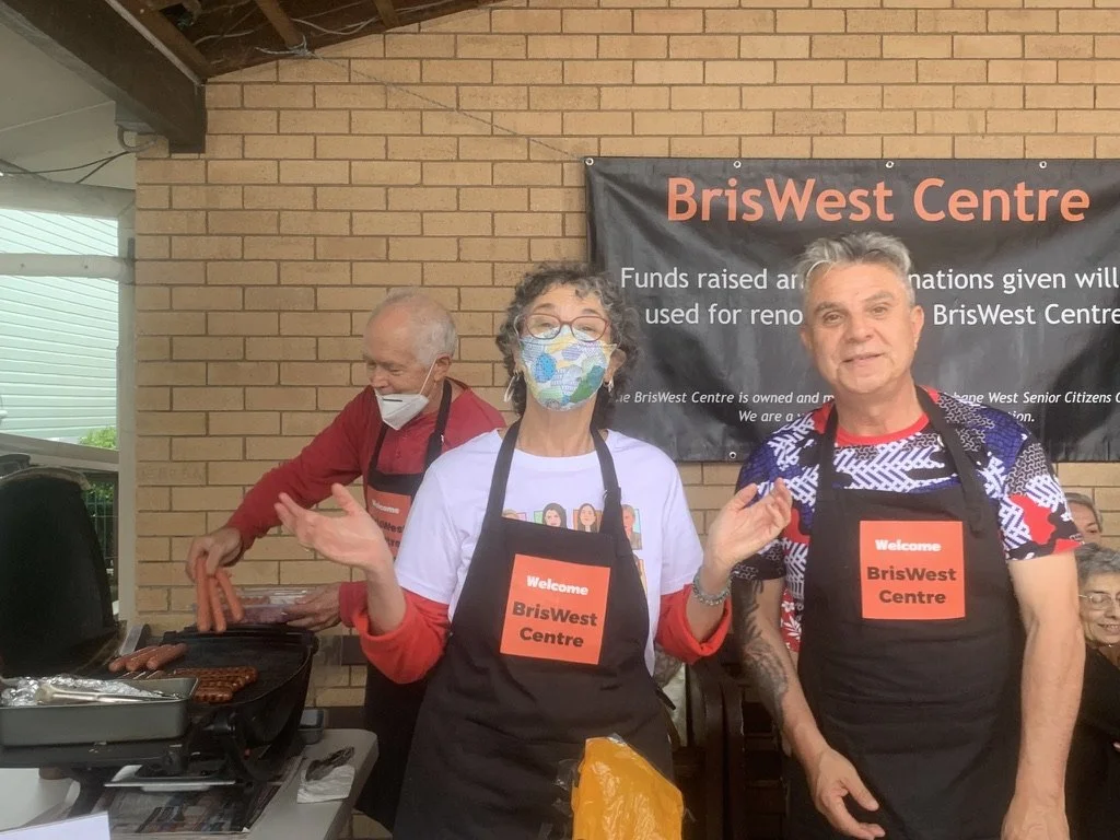 Three people standing under a brick wall with a banner that reads 'BriWes t Centre.' The two people in front are wearing welcome signs, with the woman on the left wearing a colorful face mask and a white t-shirt with cartoon characters, and the man o