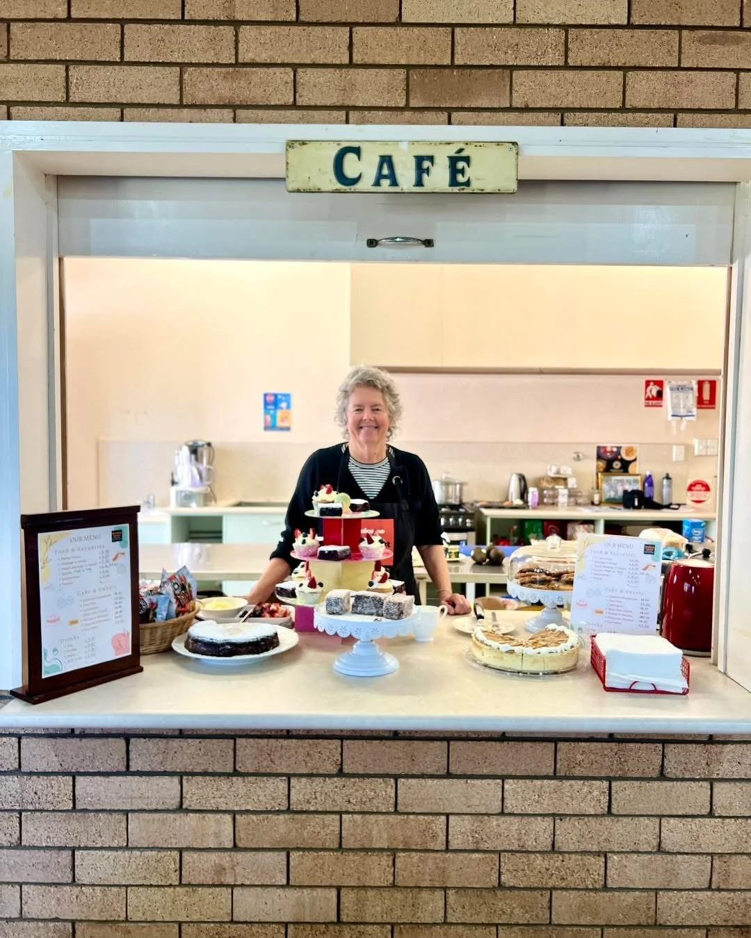 A woman smiling behind a counter at a cafe, with various desserts on display, including cakes and pastries.
