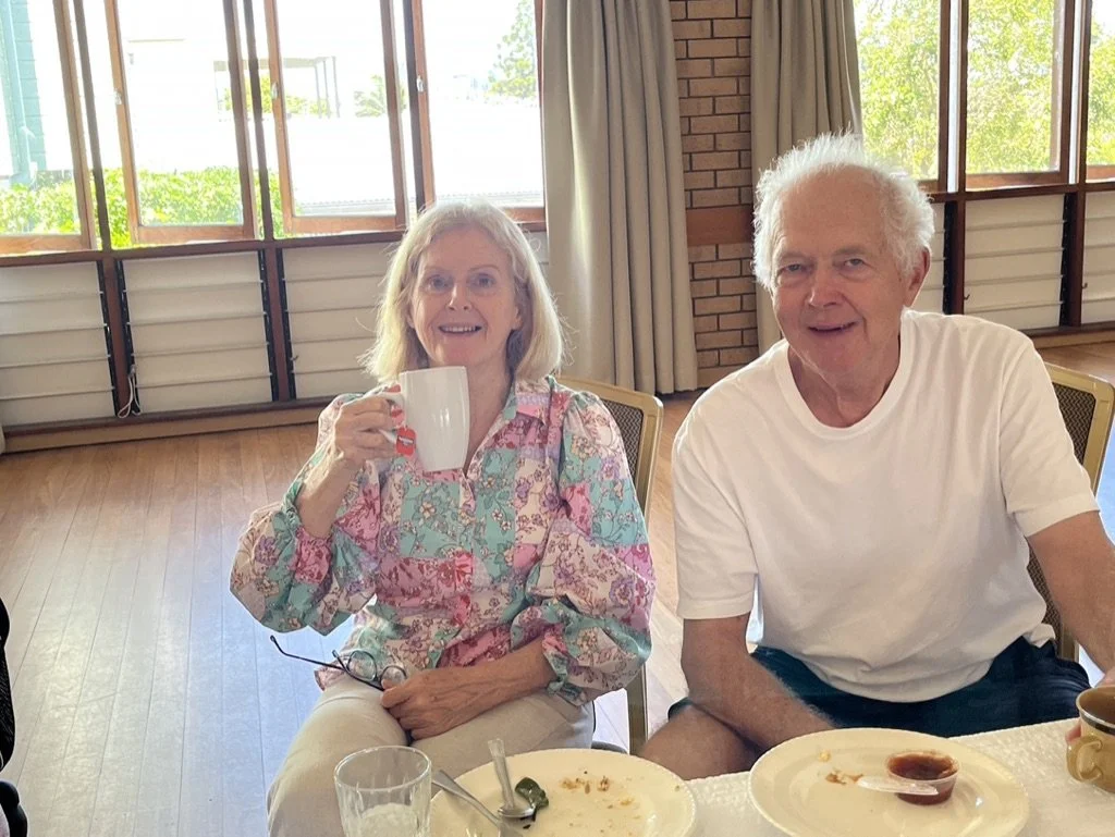 An elderly woman with gray hair wearing a colorful floral blouse, sitting at a table, holding a white mug, smiling. Next to her, an elderly man with white hair wearing a white T-shirt, also smiling. They are indoors near large windows with natural li