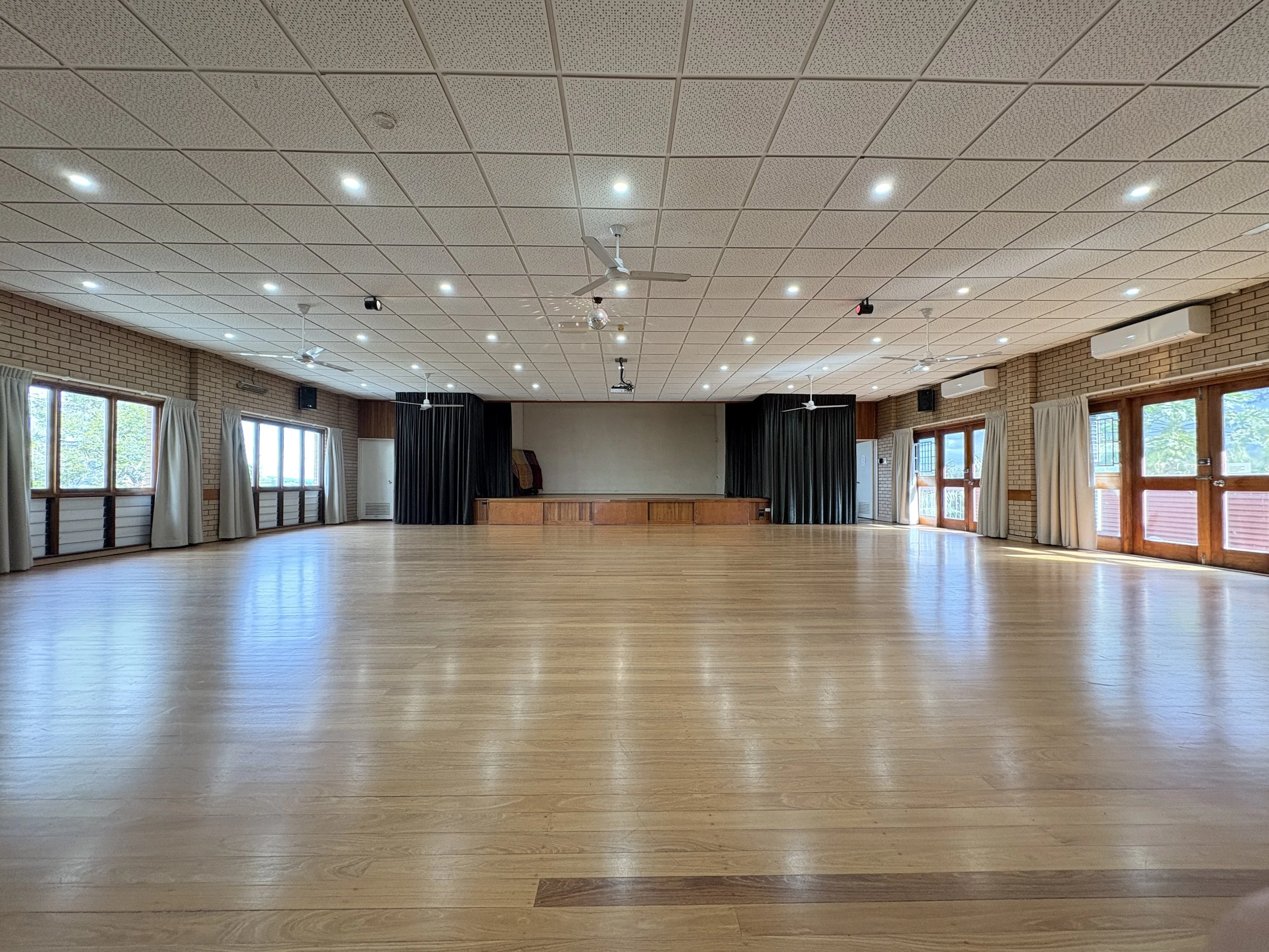 A large, empty dance hall or multipurpose room with wooden floors, large windows with curtains, and a stage at the far end with black curtains, ceiling fans, and ceiling lights.