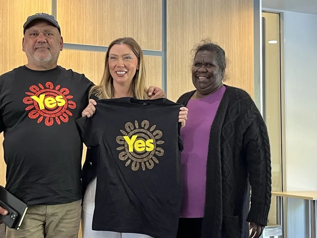 Three people smiling, holding a T-shirt with the word "Yes" and a sunburst design, standing in a room with wooden paneling.