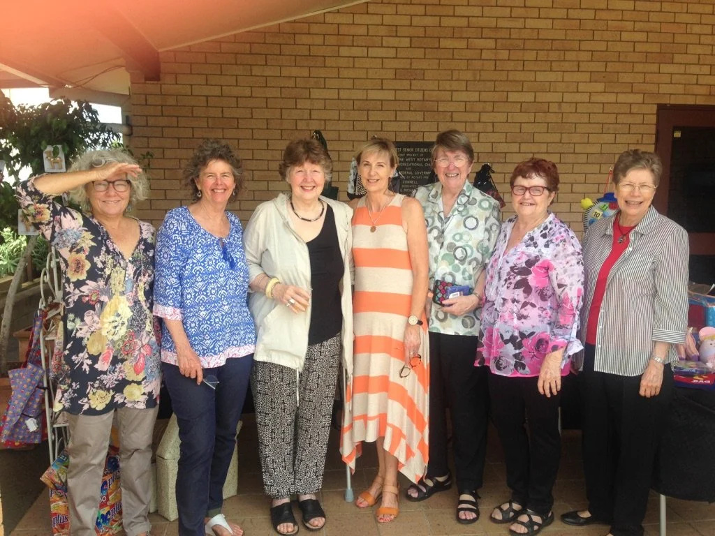 Eight women standing together outside a brick building, smiling, some holding drinks, dressed casually in colorful tops and patterned pants or skirts.