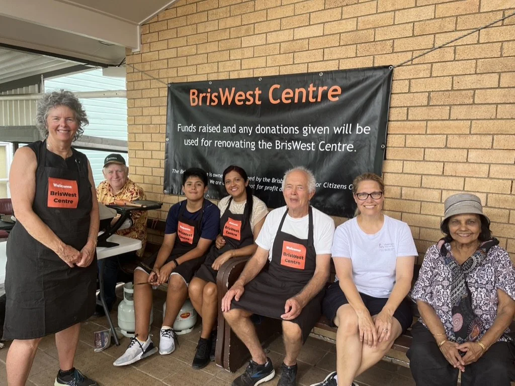 Group of six people sitting and standing in front of a banner that reads 'BrisWest Centre,' with some wearing aprons and others casual clothing, at an indoor event.