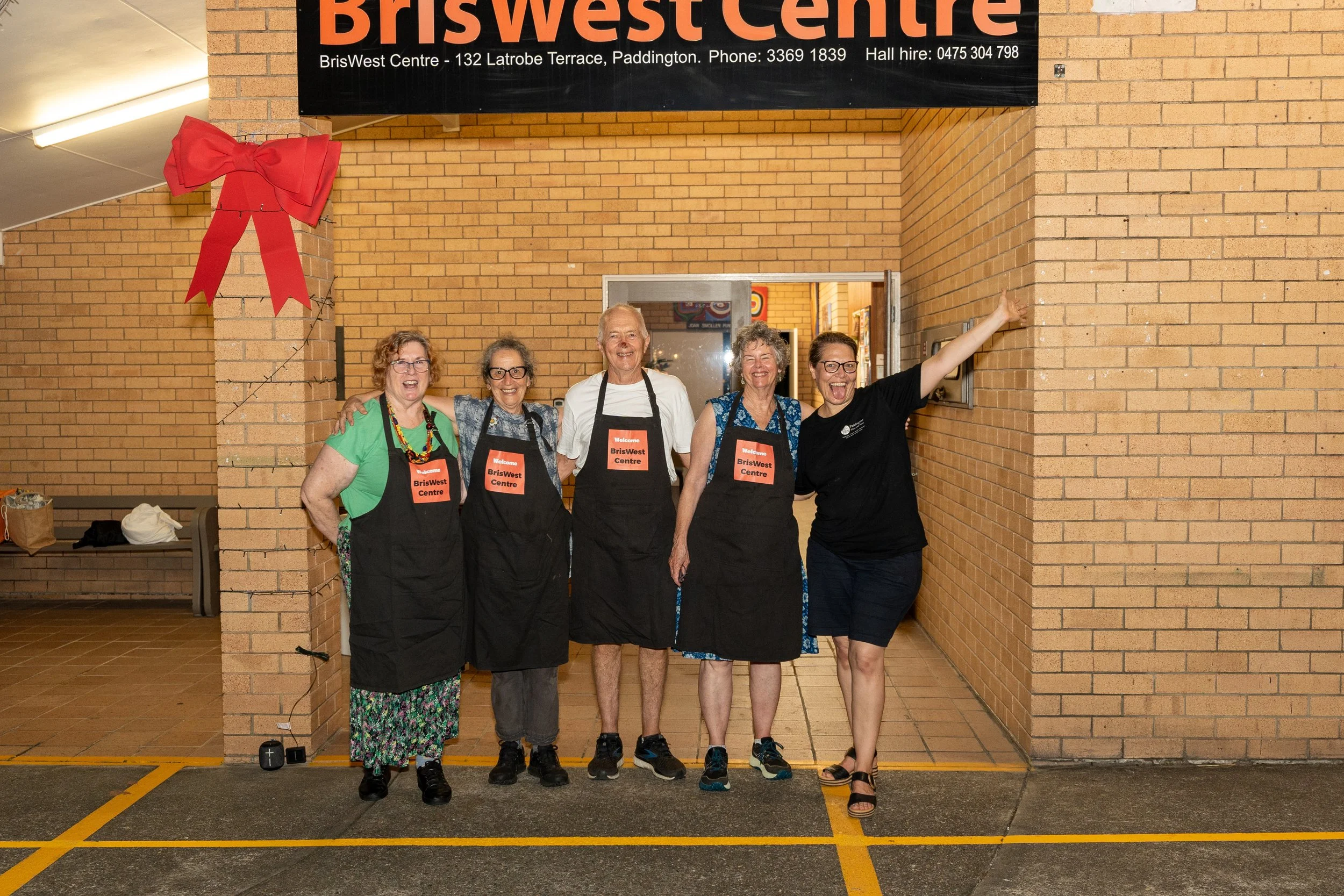 Five joyful women standing together in front of a brick wall at BrisWest Centre, all wearing black aprons with name tags. One woman on the far right is pointing towards the wall with her arm extended. The group appears happy and celebrating.