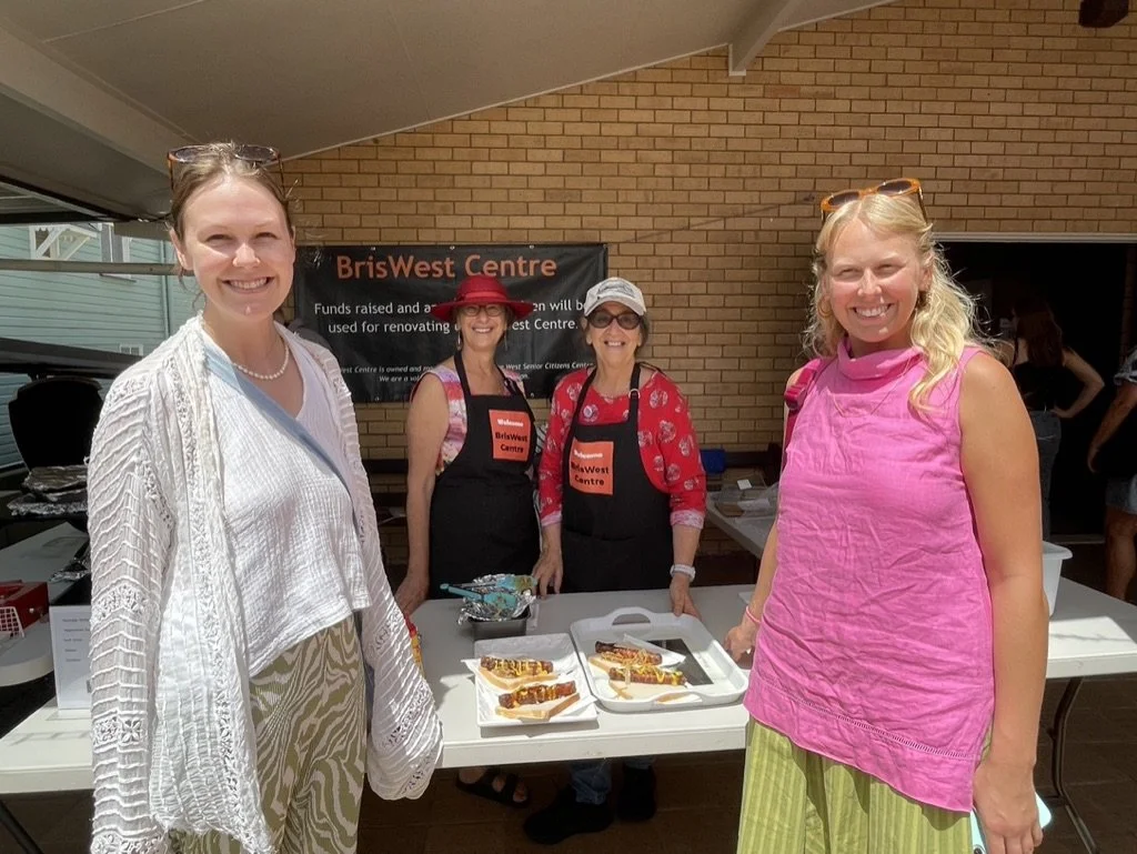 Four women smiling at a fundraiser event for BrisWest Centre, with two women wearing aprons, standing behind a table with hot dogs and condiments.