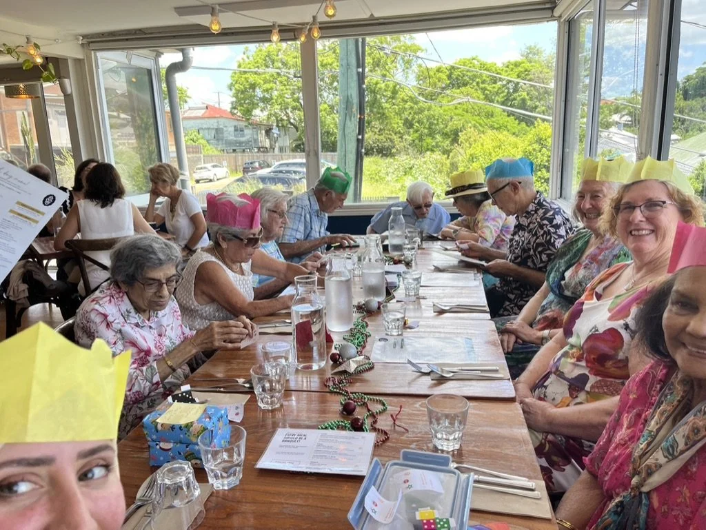 Group of elderly people celebrating at a restaurant with colorful paper crowns, Christmas decorations, and a festive setting during daytime.