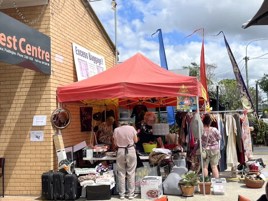 Outdoor flea market stall with clothes, accessories, and household items under a red canopy. Several women browse among hanging clothes and items on tables, with a brick wall and blue sky in the background.