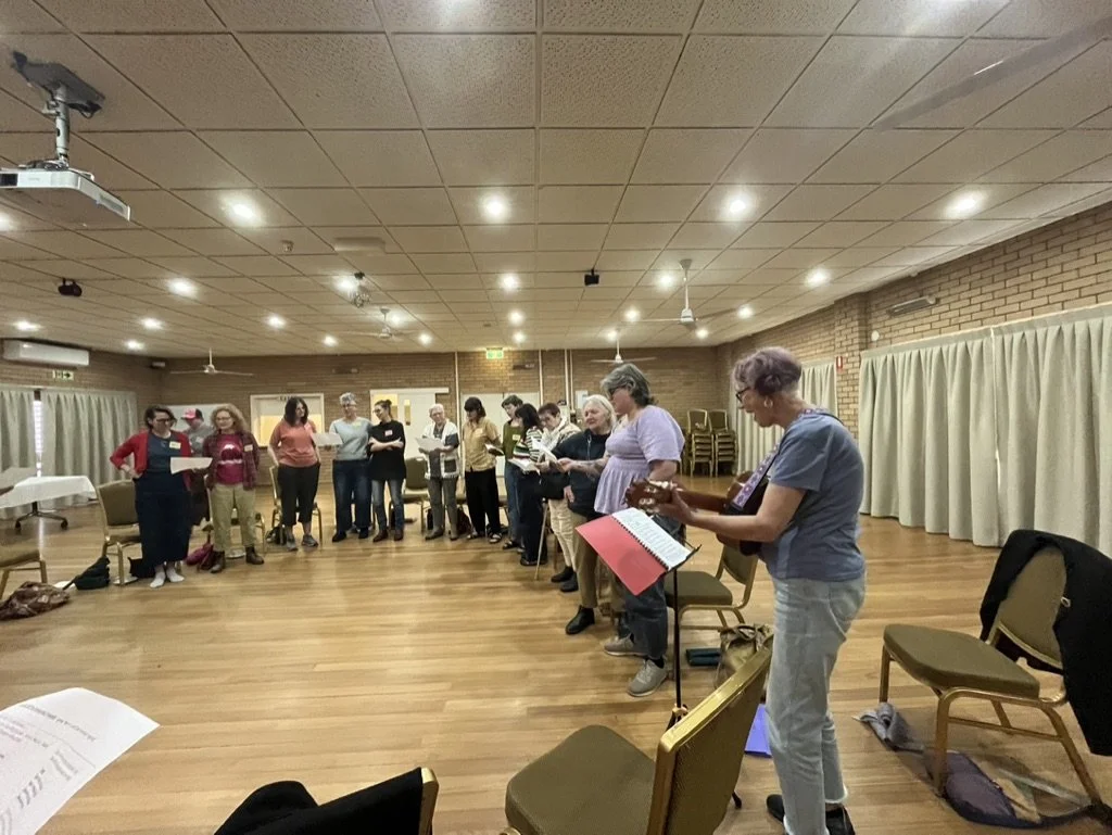 A group of people standing in a circle in a practice or rehearsal space, some holding sheets of paper, with one person playing guitar.