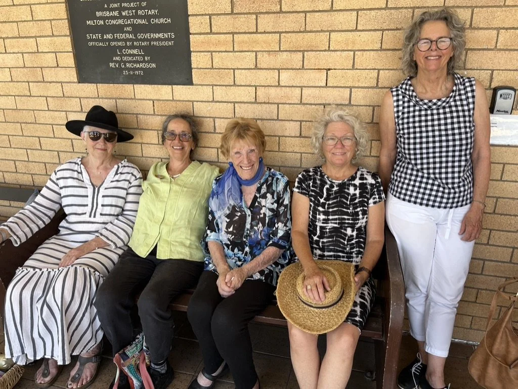 Five women sitting and standing on a wooden bench in front of a brick wall, smiling. One woman is wearing a black hat and sunglasses, another a lime green shirt, another a blue patterned shirt, and one holding a straw hat. A woman standing is wearing