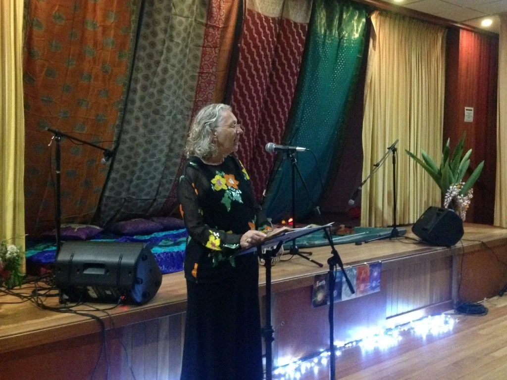 A woman with gray hair standing on a stage, speaking into a microphone, with colorful drapes and a potted plant in the background.