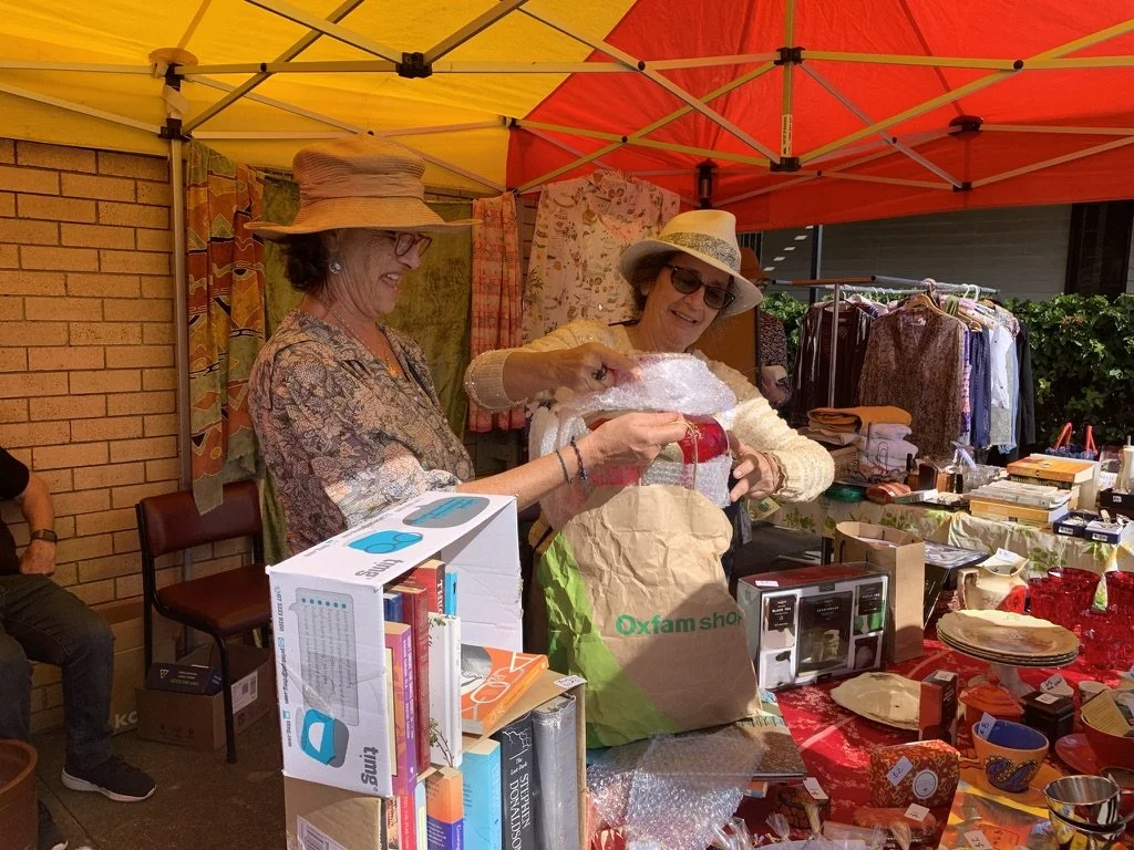 Two women wearing hats and sunglasses shopping at a table with various items, including boxes, books, and decorative objects, under a yellow and red canopy.