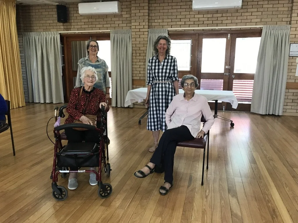Four women in a room with wooden floors and brick walls. Two women are sitting, one in a wheelchair and the other on a chair, while two women are standing behind them. All are smiling.