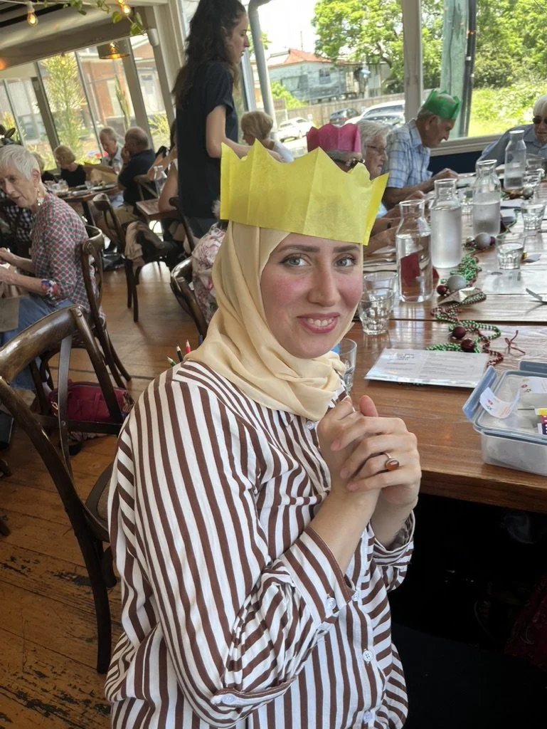 A woman wearing a beige headscarf and a striped shirt with brown and white vertical stripes, sitting at a restaurant table. She has a yellow paper crown on her head and is smiling at the camera. The table is decorated with some holiday decorations, a