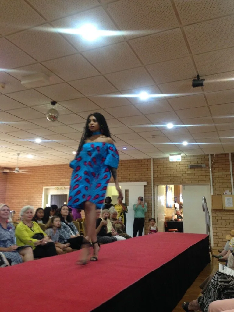 Model walking down runway at a fashion show with audience watching in a room with brick walls.