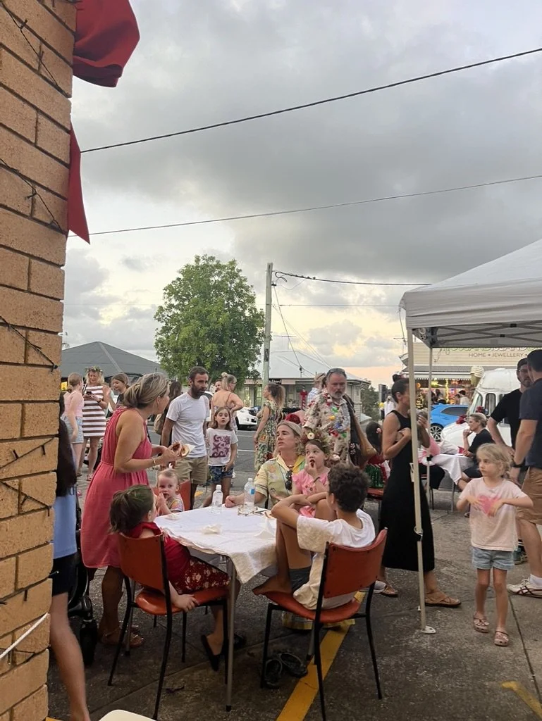 People gathered outdoors at a community event or fair, with a table in the foreground where people are sitting and talking. Some children and adults are standing around, and there is a tent with more people underneath. The sky is cloudy.