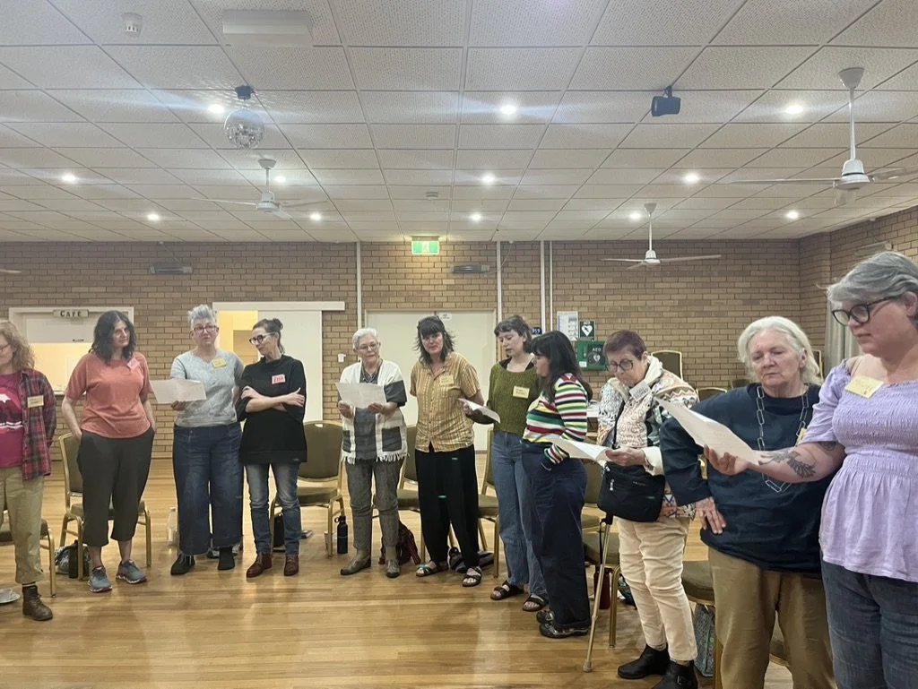 Group of women standing in a circle in a room with brick walls, some holding papers, participating in a meeting or workshop.