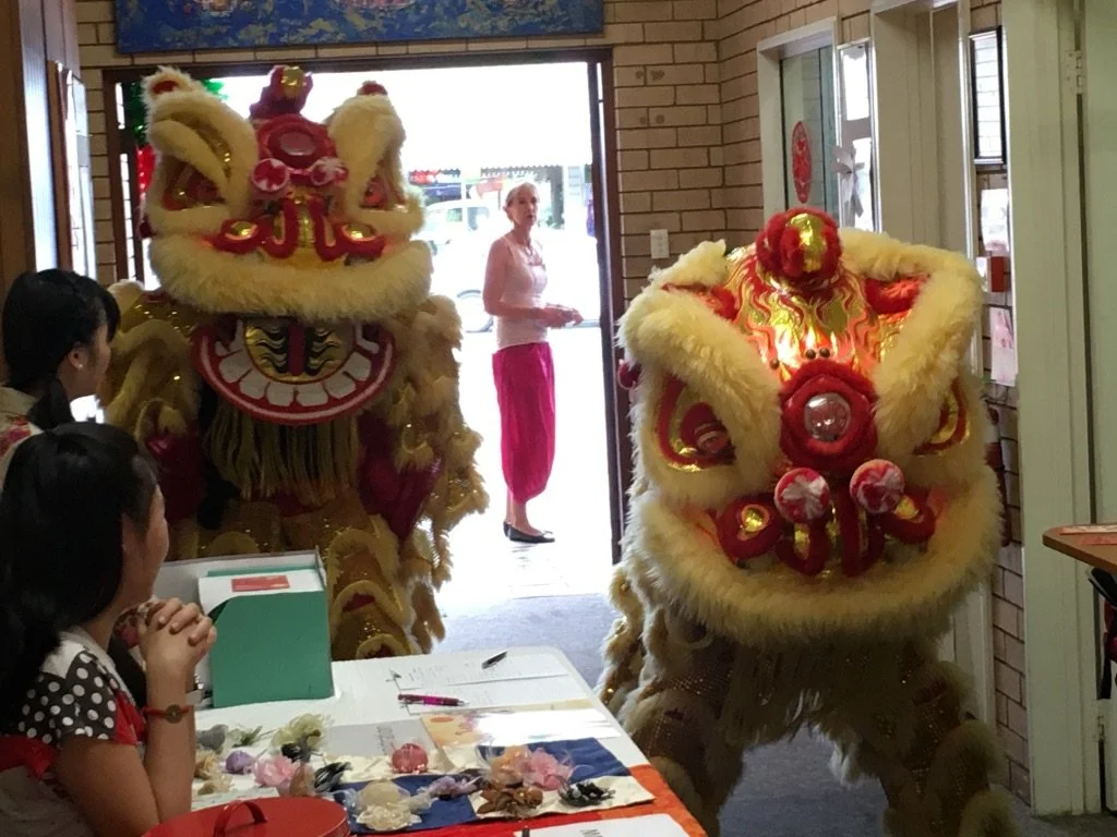 Two traditional Chinese lion dance costumes, one in yellow and the other in red, entering a room through a doorway while children and an adult watch.