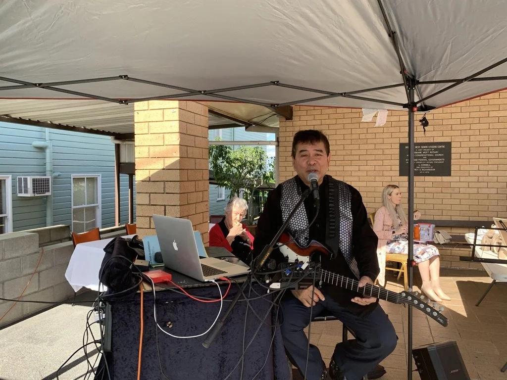 A man holding an electric guitar and singing into a microphone performing at an outdoor event under a canopy, with two women seated in the background.