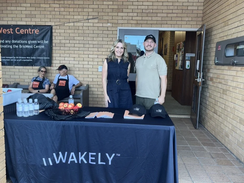 Two people standing behind a table covered with a black tablecloth with the word 'WAKELY' printed on it, at an outdoor event. The table has bottled water, apple bowls, and caps. Two elderly people sit on a bench in the background, wearing aprons. A w