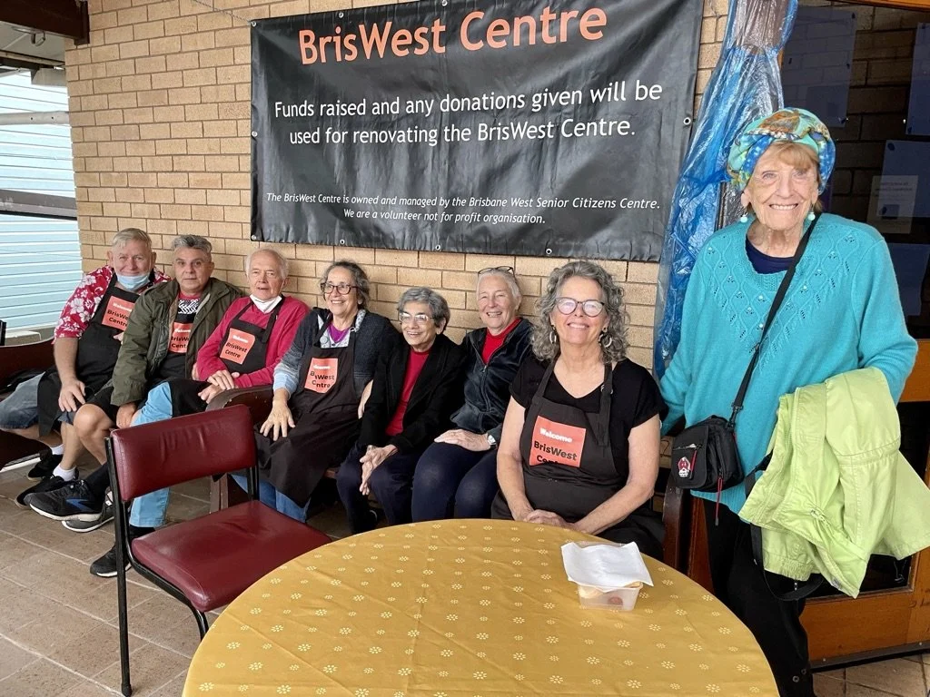 A group of smiling people sitting and standing in front of a banner that reads 'BrisWest Centre,' with some wearing aprons that say 'BrisWest Centre.' The setting appears to be a community or charity event, with a table in front of them and a brick w
