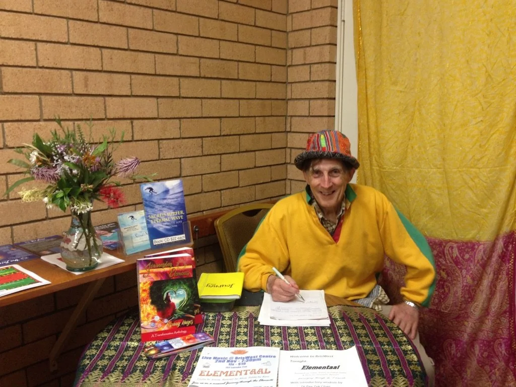 A man sitting at a table signing papers, wearing a yellow sweater and colorful hat. The table has promotional materials, books, and flyers, with a bouquet of flowers on a side table, against a brick wall and yellow curtain background.