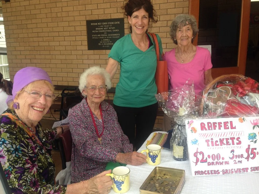 Four women, including two elderly women, are gathered around a table with raffle tickets and a sign. The women are smiling at the camera, one is seated and the others are standing. The sign indicates the raffle tickets cost $2 each, 3 for $5, and tha