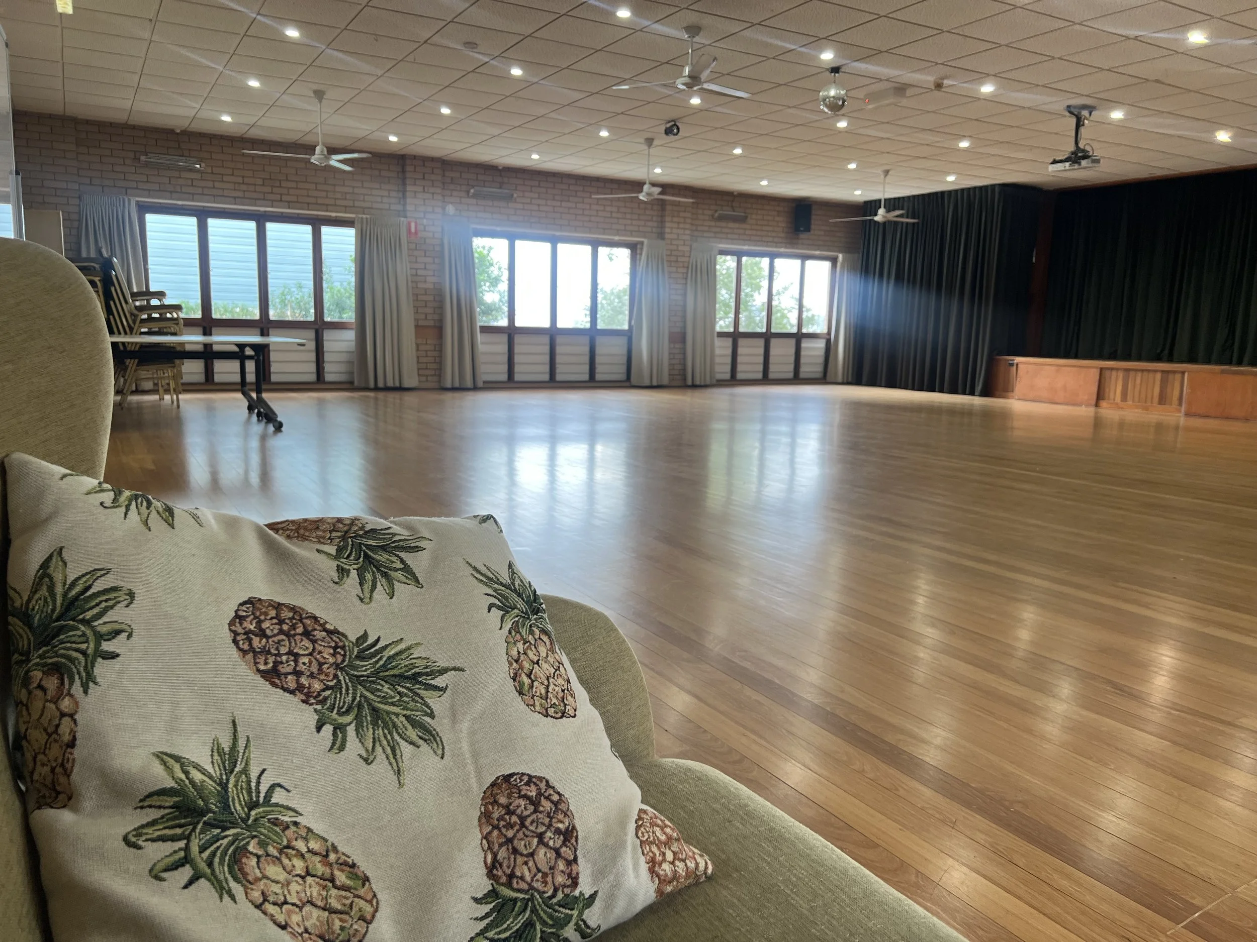 Empty multi-purpose room with wooden floors, large windows, curtains, stage with green curtains, and ceiling fans; a sofa with pineapple print cushion in the foreground.