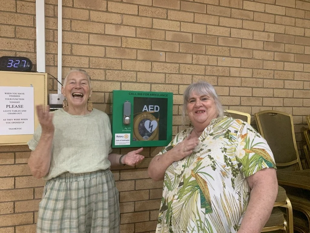 Two elderly women stand beside an automated external defibrillator (AED) mounted on a brick wall, smiling and gesturing happily.