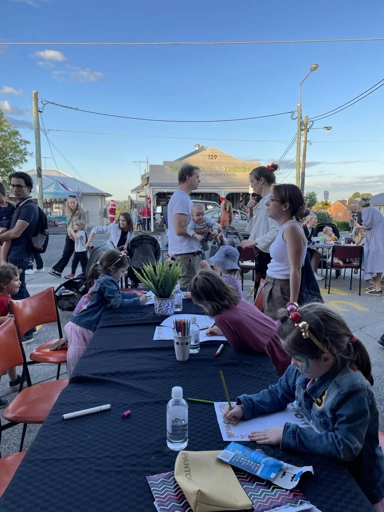 Children coloring at a table outdoors during a community event, with adults and booths in the background under clear blue skies.